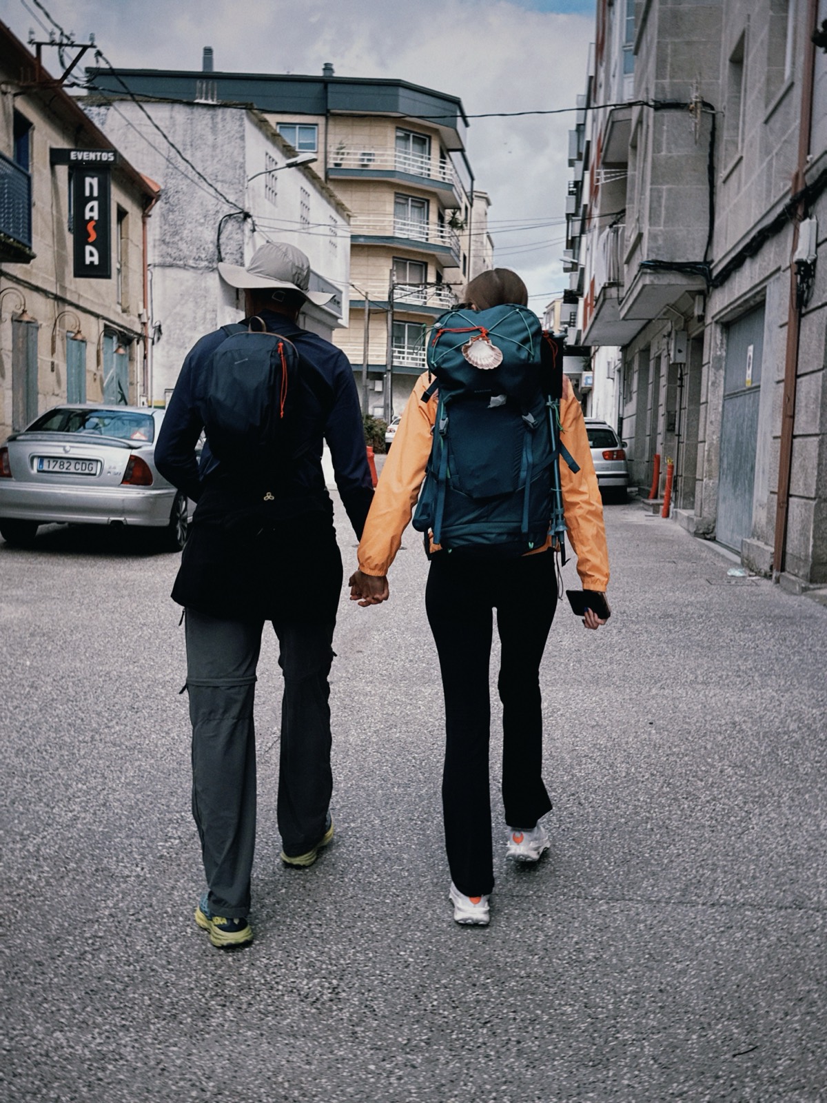 two pilgrims walking hand in hand through a galician town, scallop shell on the pack