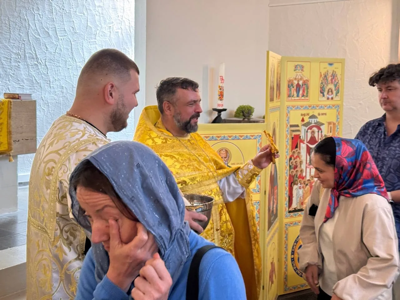 Nach der Liturgie: der Priester mit dem Kreuz spricht mit einer Frau im bunten Kopftuch, vorne eine Frau im blauen Tuch, daneben der Altardiener — eigenes Bild der Gemeinde