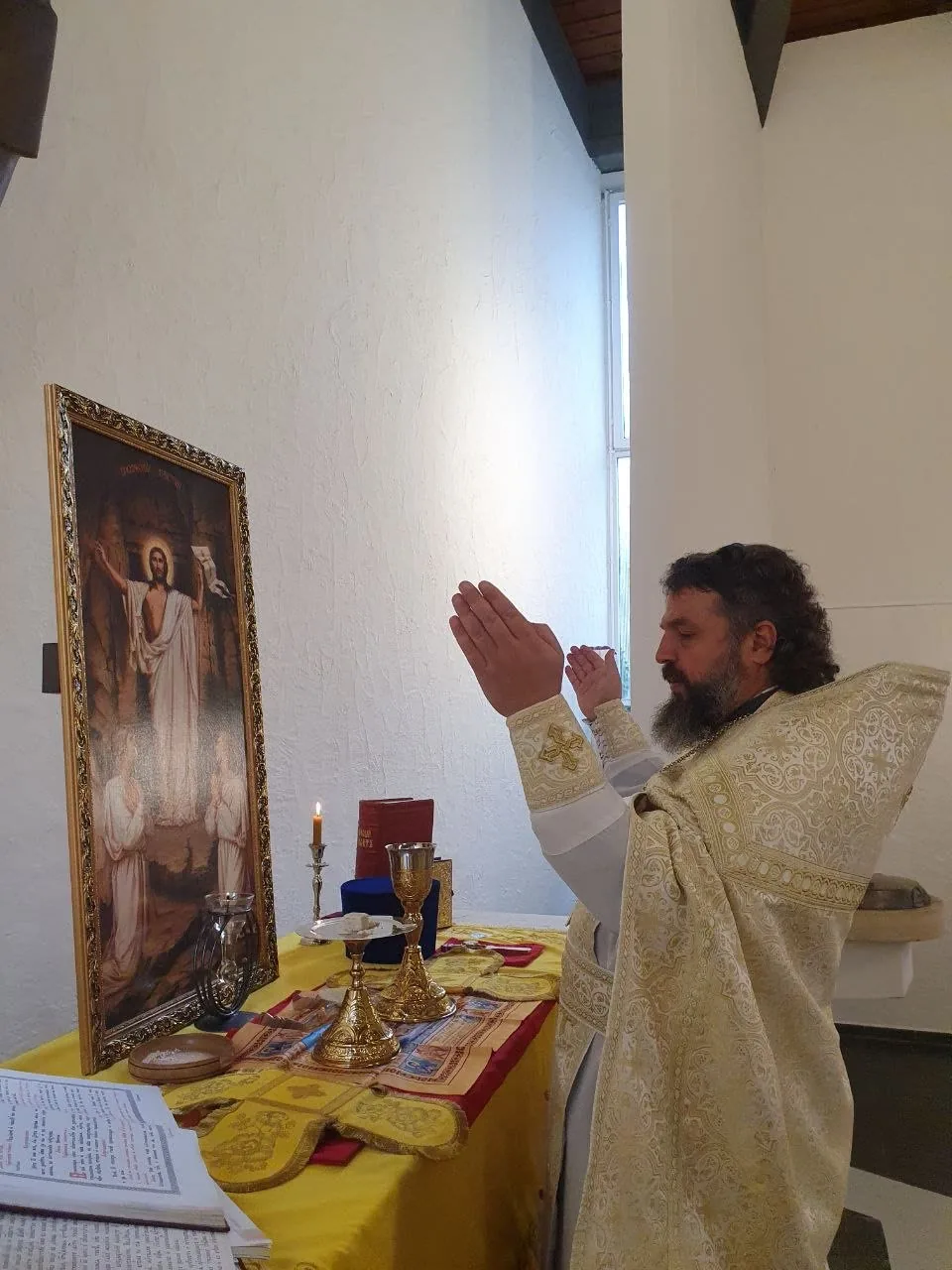 Ein Priester in goldenem Gewand betet mit erhobenen Händen vor der Ikone der Auferstehung, davor Kelch und Kerze auf dem Altar — eigenes Bild der Gemeinde