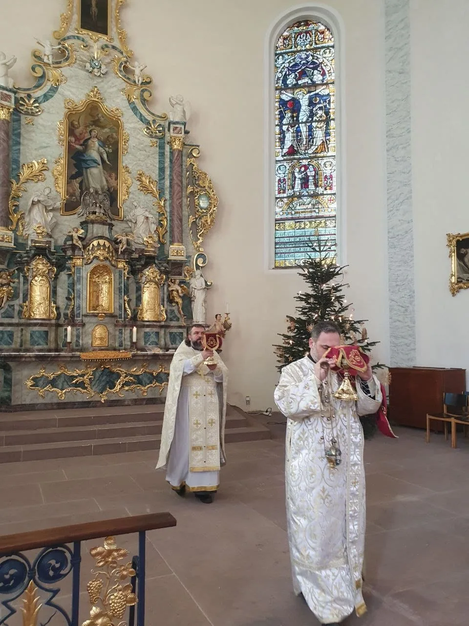 Weihnachtsliturgie: zwei Geistliche in weißen Gewändern, einer mit Weihrauchfass, vor einem barocken Altar und einem geschmückten Weihnachtsbaum