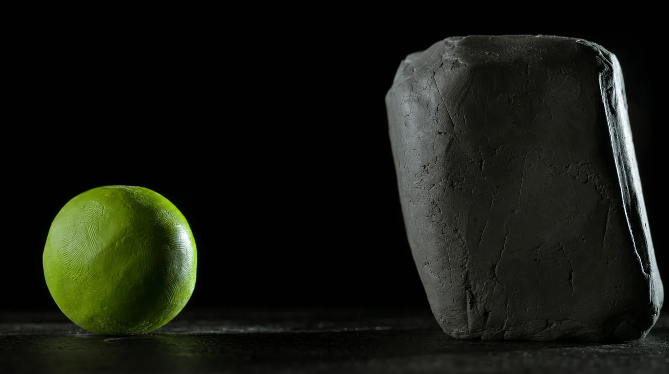 A small glossy lime-green clay sphere on the left facing a large rough grey clay block looming on the right, hard side light, black background